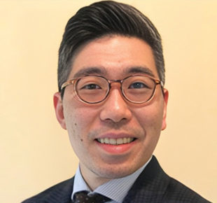 Professional headshot of an Asian man wearing glasses and dark blazer against beige background.
