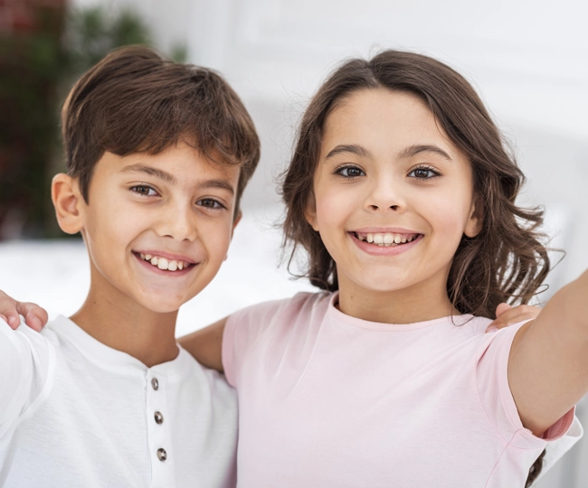 Two smiling children, a boy in white and girl in pink, posing together outdoors with snowy background.