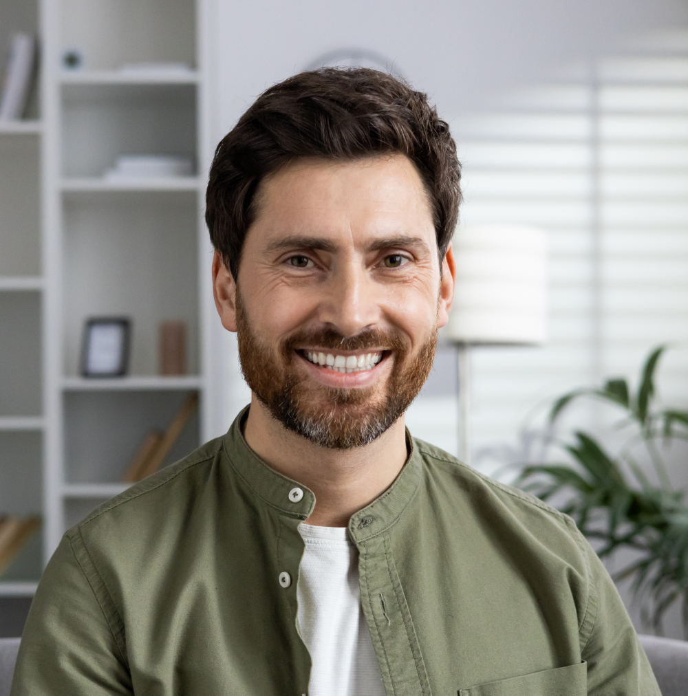Smiling man with beard wearing olive green shirt in modern office with shelving and plants.