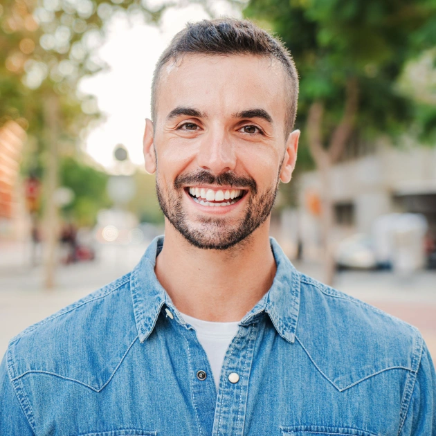 Smiling man in denim shirt standing outdoors on a tree-lined urban street.
