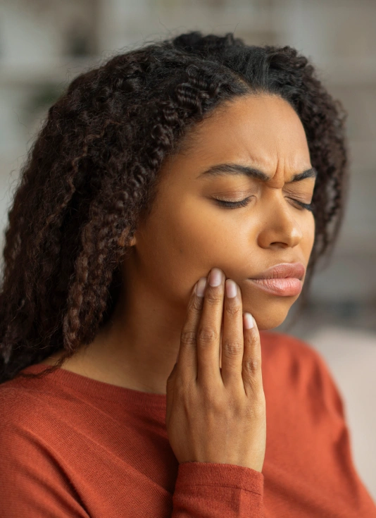 Woman with braided hair touching her cheek, appearing to have tooth pain or discomfort, wearing orange shirt.