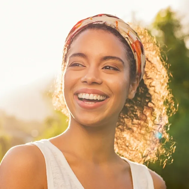 Young woman smiling outdoors in golden sunlight wearing white tank top and headband.