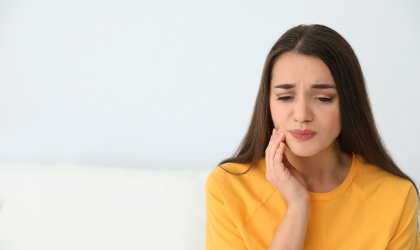 Woman in yellow shirt holding cheek, experiencing tooth pain or discomfort.