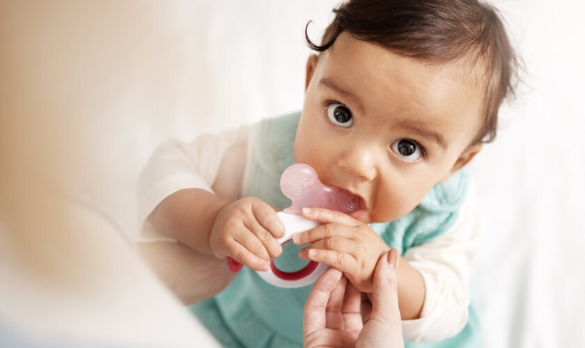 Baby chewing on pink teething ring while looking at camera with wide eyes.